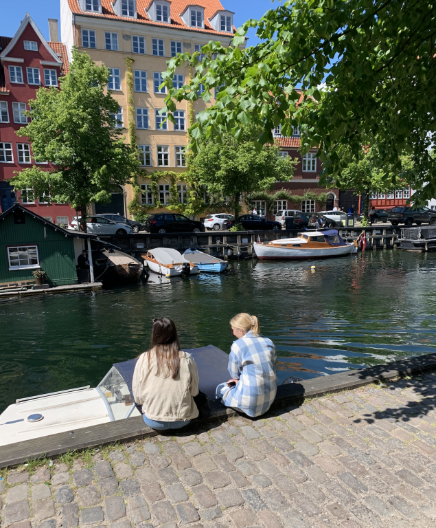 Two friends sit on the edge of a canal. Across the water, boats are docked and trees and houses line the shoreline.